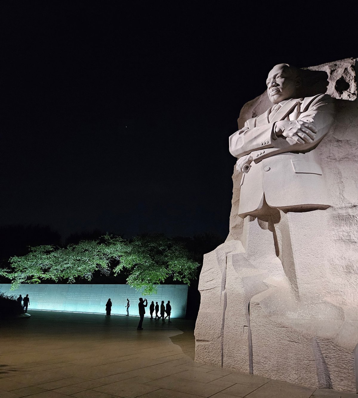 Martin Luther King Jr.'s massive statue lit up at night with a few tourists visiting. The silhouette of a tourist taking a picture of the monument can be seen against the lit wall.