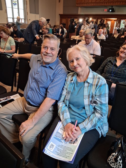 Older couple smiling at camera seated in church pews during a community gathering with many people in background