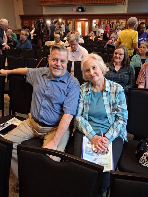 Two smiling people sitting in church pews with a crowd of worshippers gathered in a sanctuary during service