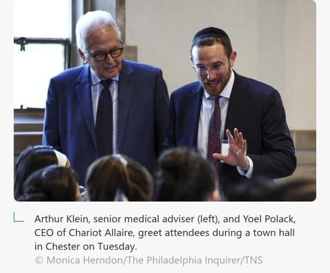 Two men in business attire greeting attendees at a town hall event, with one gesturing to the audience