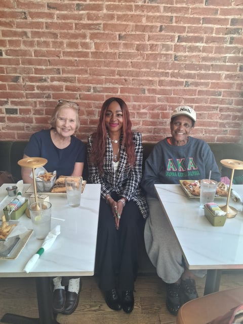 Three people seated at a dining table in front of a brick wall, enjoying a meal together
