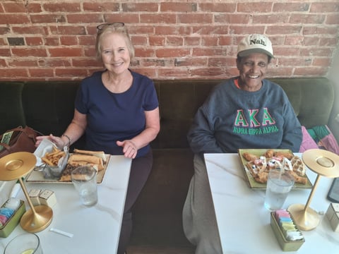 Two people sitting at a table with food in front of a red brick wall, smiling at the camera