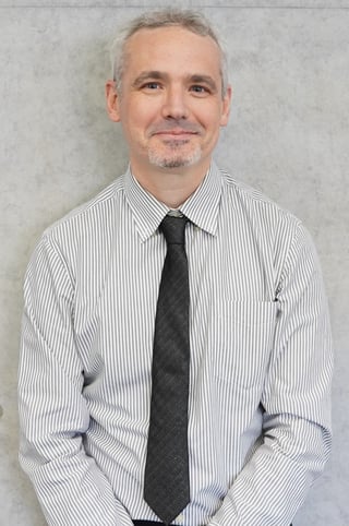 Middle-aged man with gray hair wearing a white striped dress shirt and black tie, smiling at camera in professional headshot