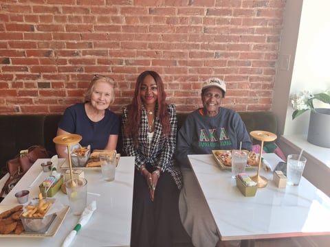 Three people sitting at a white table with food and drinks in front of a red brick wall