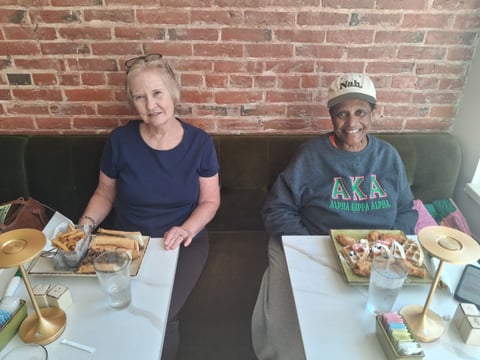 Two people sitting at a booth against a brick wall, enjoying a meal with plates of food and drinks on the table.